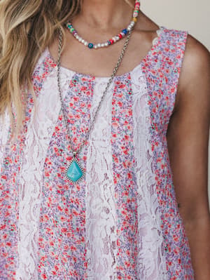 A close-up torso shot of a flowy floral dress in lilac with vertical white lace panels, layered with a colorful beaded choker and a long silver chain holding a turquoise stone pendant — the kind of outfit that feels like a farmers market and a good m