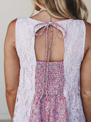 A close-up detail shot of the back of a lilac dress, showing a beautiful mix of floral lace panels and a ditsy floral print center with smocked waist detail — tied together (literally) with a dainty self-tie bow at the upper back.