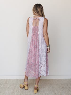 A back-facing shot of a midi dress that layers soft lilac lace panels over a ditsy floral print center, styled with yellow platform clogs and stacked boho bracelets — effortlessly romantic with a vintage-garden feel.