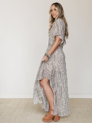 A flowy hi-low maxi dress in a soft beige botanical print styled with tan wooden clogs and layered boho bracelets — the kind of dress that looks equally at home at a farmers market or a rooftop dinner.