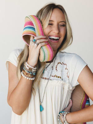 A laughing model holds a rainbow-striped crochet slide up to her face like a phone call she's been waiting for, styled in a white lace-trim top with layers of boho jewelry — the kind of shot that makes you want the shoes and the energy.