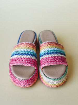 A pair of rainbow-striped crochet slide sandals shot from above against a soft sage background — the kind of shoes that make you want to book a trip somewhere warm.