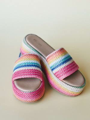 A pair of rainbow-striped crochet slide sandals with a platform sole, shot against a soft sage background — the kind of shoes that make you feel like summer is already here.