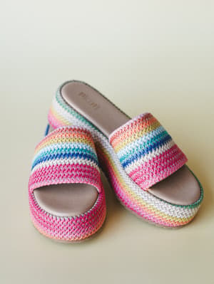 A pair of rainbow-striped crochet slide sandals with a platform sole, shot against a soft sage background — the kind of shoes that make you feel like summer is already here.