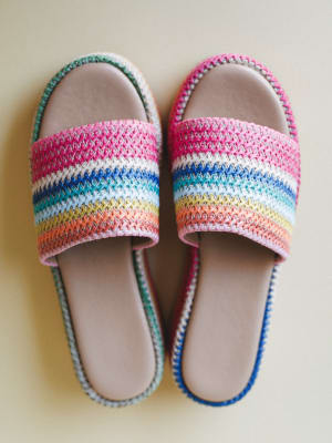 A pair of rainbow-striped crochet slide sandals laid flat on a warm cream surface — the kind of shoes that make you want to book a trip somewhere with sand.