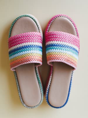 A pair of rainbow-striped crochet slide sandals laid flat on a warm cream surface — the kind of shoes that make you want to book a trip somewhere with sand.