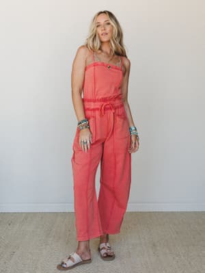 A faded coral jumpsuit with smocked waist and relaxed wide-leg silhouette, styled with turquoise jewelry and sandals against a soft neutral wall — the kind of outfit that looks effortless but clearly wasn't an accident.