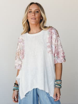 A relaxed oversized tee with a plain white knit body and sheer floral sequin sleeves in soft blush, styled with wide-leg chambray pants and stacked beaded bracelets — the kind of top that makes a casual outfit feel like it has a little secret.