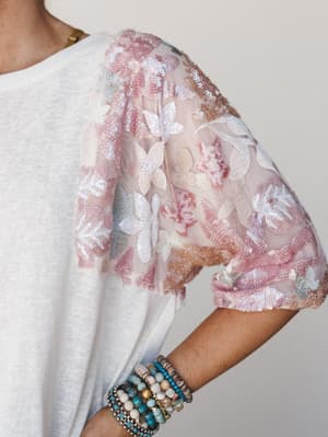 A close-up detail shot of a white tee with a dreamy, embellished puff sleeve — sheer mesh layered with floral embroidery and sequins in blush and ivory — styled with a stack of boho beaded bracelets.