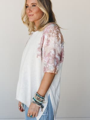 A relaxed white knit tee with sheer floral sequin sleeves in soft blush and pink catches the light beautifully — styled with stacked boho bracelets and denim for that effortless 'I just threw this on' energy that clearly took some intention.