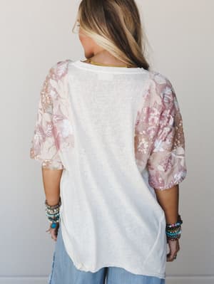 Shot from behind, a model wears a relaxed white knit tee with sheer floral sequin sleeves in blush and pale pink — the kind of top that looks effortless but clearly has something to say, styled with stacked boho bracelets and light wash denim.