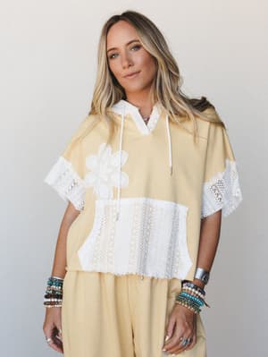 A relaxed cropped hoodie in soft butter yellow gets a boho upgrade with white crochet trim, floral appliqué, and a lace panel — worn as a matching set and stacked with an armful of beaded bracelets that feel entirely intentional.