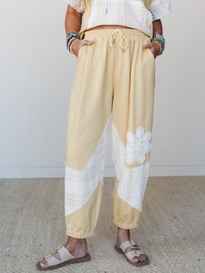 Butter yellow jogger pants with diagonal lace inset panels and a crochet flower appliqué styled with a matching cropped lace top, sandals, and stacked bead bracelets — the kind of set that makes a Tuesday feel intentional.