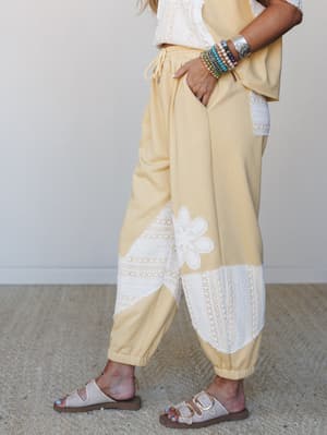 A soft butter yellow jogger with wide lace panel inserts and a daisy appliqué at the lower leg, styled with a matching lace crop top, stacked bracelets, and slide sandals — the kind of set that makes a coffee run feel intentional.