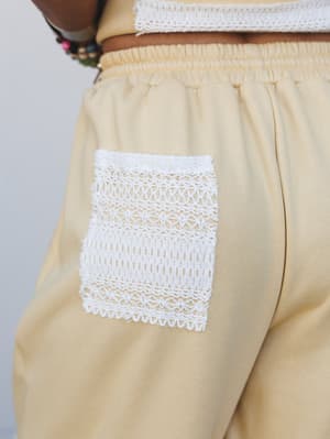 A close-up detail shot of butter cream pants with a crochet lace patch pocket, paired with a matching top trimmed in the same white lace — the kind of set that feels handcrafted without trying too hard.