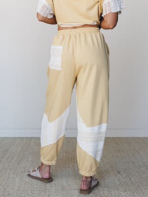 Shot from behind, a model wears a butter yellow jogger and matching cropped top set with bold crochet lace panel inserts at the lower legs and back pocket — relaxed, boho-meets-cozy energy with a warm, soft feel.