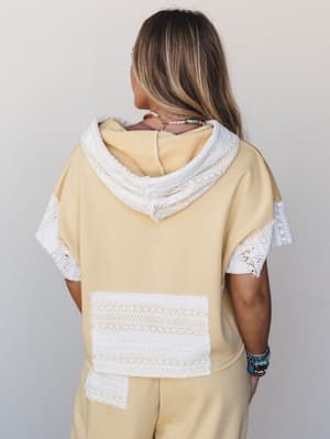 A back view of a butter cream short-sleeve hoodie with white crochet lace trim detailing on the hood, sleeves, and back pocket, styled as a matching set with coordinating wide-leg pants and stacked boho jewelry.