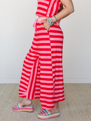 A side-profile shot of a bold red and pink striped matching set — wide-leg pants with a tie waist and a coordinating striped top — styled with rainbow platform sandals and stacked turquoise bracelets for a playful, color-saturated look.