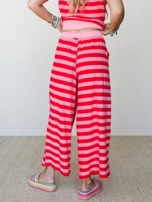 A back-view shot of wide-leg striped pants in bold red and pink, styled as a matching set with a cropped top, rainbow platform slides, and stacked turquoise bracelets — the kind of outfit that's clearly having a good time.