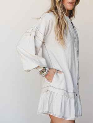 A breezy oatmeal linen mini dress with contrast topstitching and a flounced hem, styled with a stack of boho beaded bracelets — the kind of outfit that looks effortless but clearly wasn't an accident.