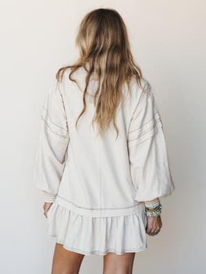 Shot from the back, a relaxed mini dress in oatmeal linen moves softly against a neutral studio wall — the billowy balloon sleeves, contrast stitching, and ruffle hem giving it that effortless, lived-in boho feel.