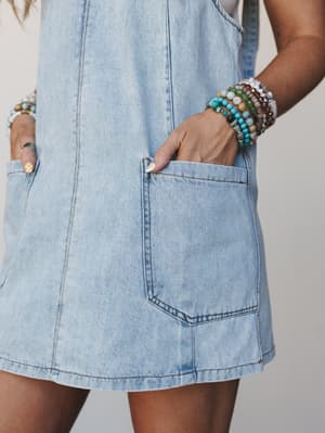 A close-up mid-shot of a light denim overall mini dress with front patch pockets, styled with a stack of colorful beaded bracelets tucked casually into the pocket — relaxed, sun-worn, and ready for wherever the day goes.