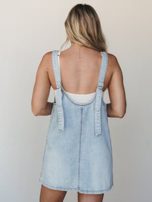 A back-view shot of a light wash denim overall mini dress layered over a white ribbed tank, styled simply against a soft neutral studio wall — effortless and a little nostalgic.