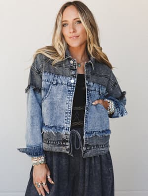 A two-tone acid wash denim jacket with fringe trim detailing worn open over a black crochet top and charcoal midi skirt, styled with stacked beaded bracelets and layered rings for an effortlessly boho, lived-in look.