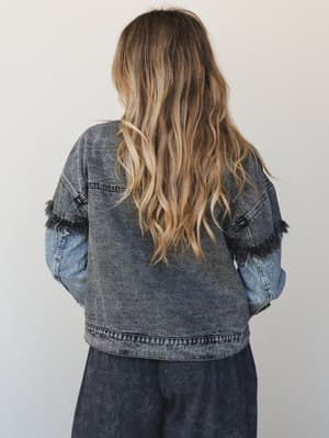 Shot from behind, this two-tone denim jacket pairs an acid-washed charcoal body with lighter blue sleeves and fringe trim detailing at the arms — worn over dark wide-leg pants for an effortlessly cool, layered look.