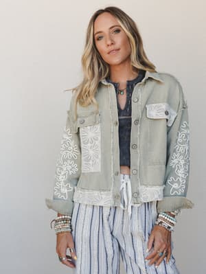 A relaxed, layered look built around a sage canvas jacket covered in white embroidery and lace patchwork — styled over a charcoal crop top and striped wide-leg pants, with stacked beaded bracelets and a turquoise necklace pulling the whole boho story