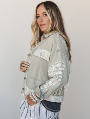 A sage denim jacket with white floral embroidery on the sleeve and lace trim details, styled over a navy stripe pant — the kind of layering piece that makes a casual outfit feel intentional and a little collected.