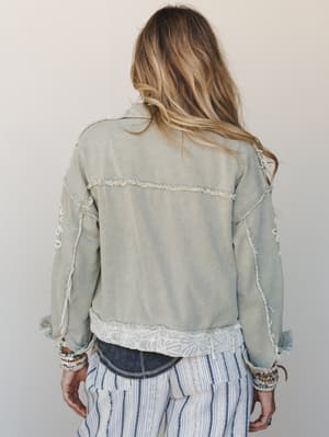 Shot from behind, this washed sage denim jacket layers effortlessly over striped wide-leg pants, its raw frayed seams and lace hem trim giving it that perfectly undone, lived-in boho energy.