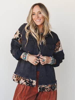 A smiling model layers a washed navy denim jacket with tapestry floral patchwork panels and frayed trim over a rust corduroy skirt, styled with stacked beaded bracelets and silver rings for a lived-in boho feel.