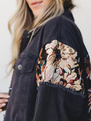A close-up of a washed navy denim jacket with a bold tapestry panel across the sleeve — the kind of jacket that looks like it has a whole personality before you even put it on.