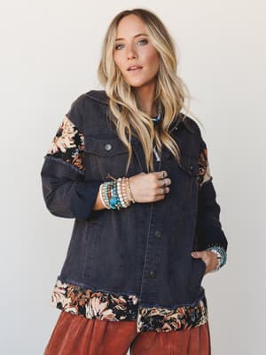 A washed navy denim jacket with floral tapestry patchwork panels at the sleeves and hem, styled over a rust corduroy bottom and layered with stacked boho bracelets — the kind of jacket that makes a whole outfit feel like a personal collection.
