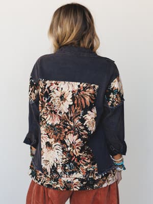 A back-view shot of a mixed-media denim jacket in washed navy, with a bold tapestry floral panel covering the back yoke and lower body, styled over rust corduroy pants with stacked beaded bracelets peeking out at the wrists.