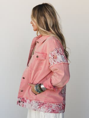 A side-profile shot of a pink washed denim jacket with floral tapestry panel inserts at the hem and sleeves, styled over a white lace dress and stacked with chunky mixed bracelets — the kind of piece that makes a simple outfit feel like a whole perso