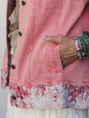 A close-up detail shot of a washed pink denim jacket with a floral tapestry hem band and frayed trim, styled over a lace layer — her hand tucked into the pocket and loaded with stacked stone bracelets tells the whole story.
