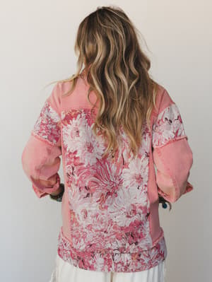 A back-view shot of a pink denim jacket with tapestry floral panels covering the back, sleeves, and hem — the kind of piece that looks like it wandered out of a vintage shop and never looked back.