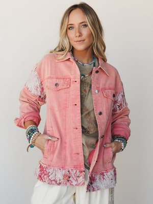 A pink washed denim jacket with floral patchwork panel inserts at the sleeves and hem worn open over a graphic tee, styled with white wide-leg pants and stacked boho bracelets — the kind of jacket that turns a lazy outfit into a whole moment.