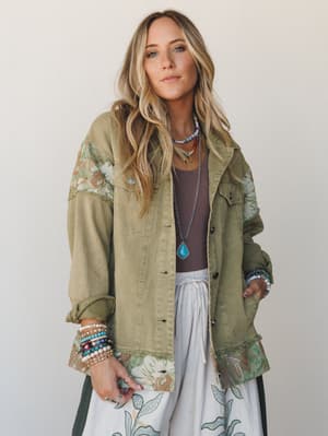 An oversized olive utility jacket with floral tapestry panels at the shoulders, cuffs, and hem is styled over a mocha tank and white wide-leg pants, layered with turquoise jewelry and stacked beaded bracelets for a free-spirited, collected look.