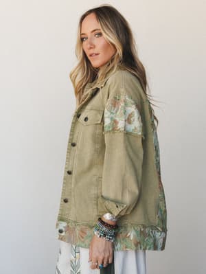 A relaxed oversized denim jacket in washed sage olive gets a boho upgrade with floral tapestry panels at the shoulders and hem — worn over a flowy white skirt with stacked turquoise and wood bead bracelets, the whole look feels like something you'd t