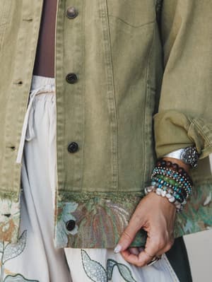 A close-up of a sage olive utility jacket with a tapestry hem detail, layered over a white floral skirt, finished with a stack of beaded stone bracelets and a silver cuff — the kind of outfit that looks like it came together effortlessly but definite