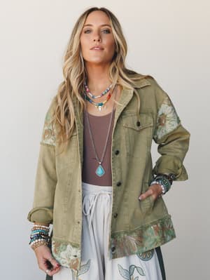 A relaxed, well-traveled vibe — an olive utility jacket with floral tapestry patchwork at the shoulders, cuffs, and hem, worn open over a mauve tank and white drawstring pants, layered with turquoise jewelry and stacked beaded bracelets.