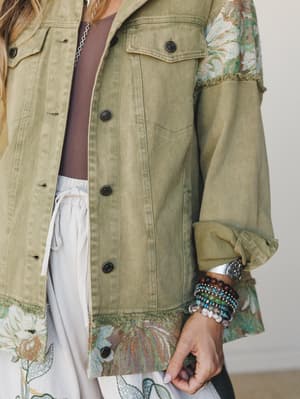 A close-up of a sage olive denim jacket with tapestry-panel sleeves and frayed hem detailing, layered over a white drawstring skirt — styled with a stack of beaded bracelets and a silver cuff that feel as intentional as the outfit itself.