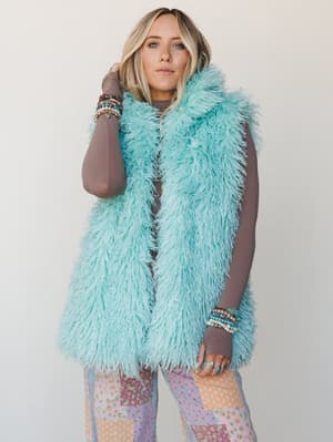 A mint faux fur shaggy vest layered over a taupe long-sleeve top with patchwork floral wide-leg pants — the whole look is playful and free-spirited, finished with an armful of stacked bracelets and rings that feel totally intentional.