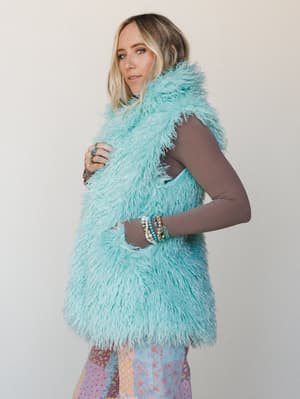 A shaggy mint faux fur vest layered over a taupe long-sleeve top, styled with a patchwork floral skirt and a stack of beaded bracelets — playful and free-spirited with a vintage-boho heart.