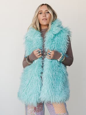 A mint faux fur shaggy vest layered over a long-sleeve mocha top and patchwork floral pants, styled with stacked bracelets and silver rings — the kind of outfit that has a personality before you even walk in the room.
