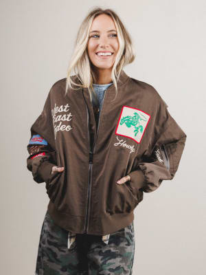 Smiling woman wearing a brown jacket with patches and text, standing against a plain background