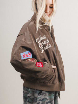 Woman wearing a brown jacket with patches and text, standing against a plain background